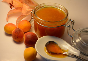 Jam in a glass jar, spoon in a plate, apricots and a flower of an orange lily in the background