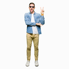Handsome man wearing fashion sunglasses smiling with happy face winking at the camera doing victory sign. Number two.