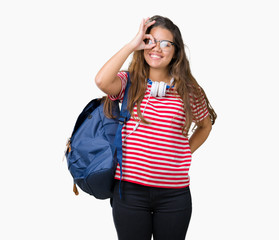 Obraz na płótnie Canvas Young beautiful brunette student woman wearing headphones and backpack over isolated background doing ok gesture with hand smiling, eye looking through fingers with happy face.