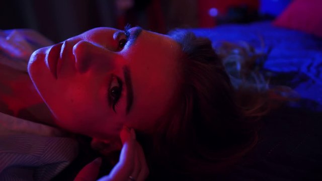 Close up view of gorgeous young woman lying in bed and playfully looking straight to camera in neon lights. Female portrait. Seduction concept. Natural beauty, hot, facial expression