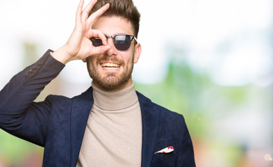 Young handsome elegant man wearing sunglasses doing ok gesture with hand smiling, eye looking through fingers with happy face.