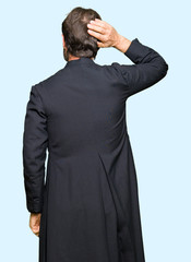 Middle age priest man wearing catholic robe Backwards thinking about doubt with hand on head