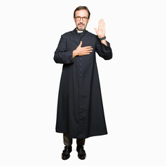 Middle age priest man wearing catholic robe Swearing with hand on chest and open palm, making a...