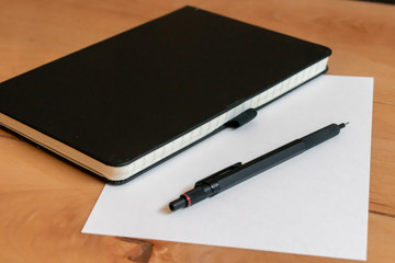 Black leather notepad and modern metallic mechanical pencil with a white sheet of paper on a bright wooden desk