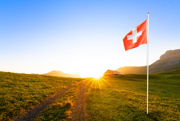 swiss chalet or farm in mountain landscape at sunrise with sun star and swiss flag waving in the foreground 