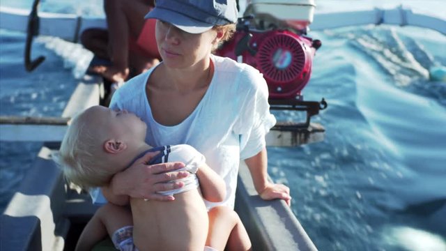 Caucasian mother is holding two-years old sleeping son while sailing in traditional indonesian canoe, motor jukung, surching for dolphins in the morning. Bali, Indonesia