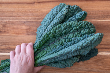Woman’s hand holding a bunch of dark green, crinkly, Lacinato Kale leaves, on a wood cutting board