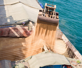 Loading wheat on cargo ship with crane and bucket. Shipment from a merchant ship to a small ship. Grapple crane fill wheat into cargo ship.