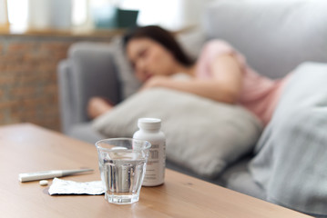 Obraz na płótnie Canvas Unwell condition of Asian women sleeping on sofa after checking temperature and taking medicines. Focus on glass of water put on the table. She has a fever and feeling bad. Sickness concept.