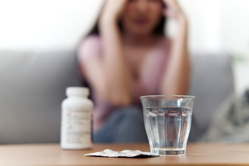 Unwell condition of stressful Asian women having headache sitting on sofa at home. Focus on glass of water put on the table. She has a fever and feeling bad. Medical sickness and illness concept.