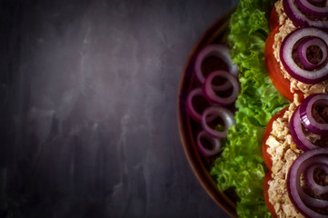 Obraz na płótnie Canvas Tuna sandwich on dark background. Selective focus. Clay plate. Fish, lettuce, red onion, tomato, bread. Free space.