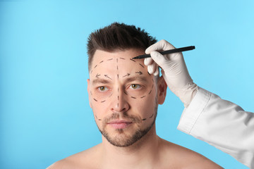 Doctor drawing marks on man's face for cosmetic surgery operation against blue background