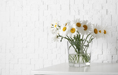 Beautiful tender chamomile flowers in vase on wooden commode near white textured wall, space for text