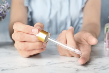 Woman holding dropper with lavender essential oil at marble table, closeup