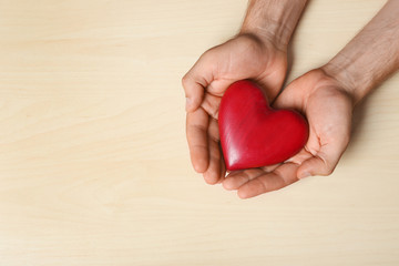 Young man holding red heart on light wooden background, top view with space for text. Donation...