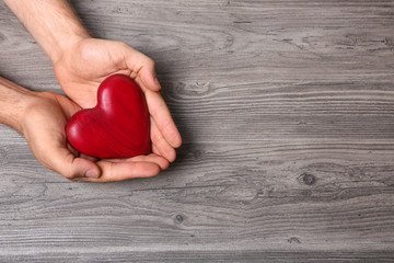 Young man holding red heart on grey wooden background, top view with space for text. Donation...