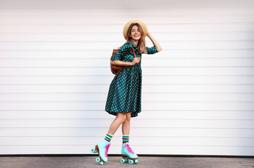 Happy stylish young woman with vintage roller skates, hat and backpack near white garage door on...