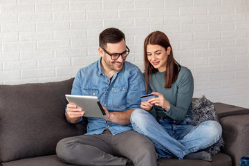 Young couple sitting on sofa with tablet and credit card on hand spending time on online shopping from room