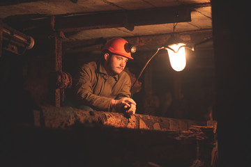 Fototapeta premium A young guy in a protective suit and helmet sits in a tunnel with a burning scrapbook. Miner in mine