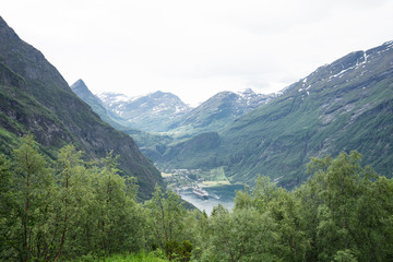 Fototapeta na wymiar Panoramic view from the high mountains down to the Geiranger Fjord (UNESCO World Heritage Site). Three huge sea cruisers which look tiny like matchboxes in the scenic bay. Green foliage framing.