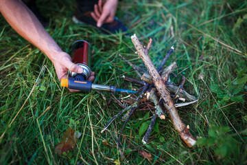 The use of a gas burner with a cylinder to ignite the fire. A man's hand holds a burner with a red cylinder and ignites the wood.