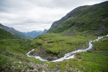 Panoramic view at the river, waterfall and peaks of the mountain range next to Geiranger village and fjord on a cloudy and humid summer day in Norway. Road E 63. Scandinavia, Europe