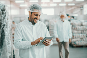 Smiling Caucasian supervisor in sterile uniform standing in food factory with tablet in hands and...