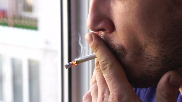 A young handsome caucasian man with a stubble on his face smokes a cigarette on the balcony and blow up smoke, close-up, slow motion
