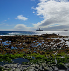 Fototapeta na wymiar Colorful rocky coast at low tide with oil rigs in the bay and blue sky with clouds 