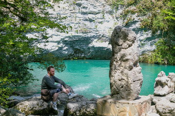 Young man sits on a stone on the shore of a mountain lake. In front of him there is a big megalith...