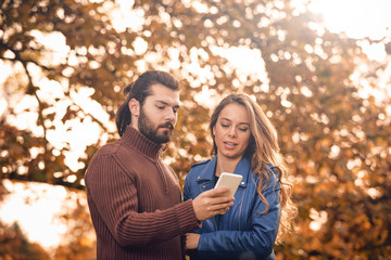 Young couple using cellphone in autumn colored park.