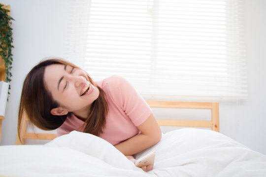 Young Asian woman sitting on the bed bursting out laughing when she hearing something interesting.