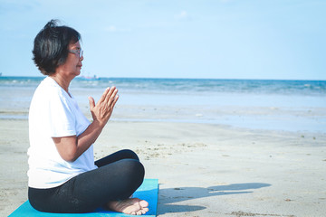 An old Asian woman wearing a white shirt is doing yoga sitting on the beach.