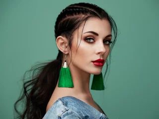 Brünettes Mädchen mit perfektem Make-up. Schöne vorbildliche Frau mit lockiger Frisur. Pflege- und Schönheitsprodukte für das Haar. Dame mit geflochtenem Haar. Modell mit Schmuck. Türkisfarbener Hintergrund © Oleg Gekman