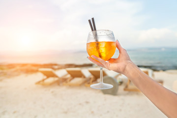 Woman holding Delicious classic iced aperol spritz cocktail with ice cubes on a hot tropical beach in summer sunshine
