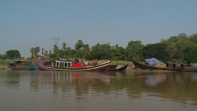 A daylight medium shot from inside a moving passenger boat traveling in the murky river water passing by the riverside where a few motorboats and pump boats are parked.