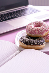 Close-up. Fresh tasty strawberry pink and chokolate donuts, in icing and sprinkling on a plate and an open notebook and laptop on a pink background. Coffee break. Vertical