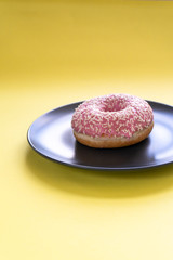 Fresh tasty strawberry donut, in icing and sprinkling on a plate on a yellow background.  Vertical