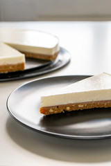 A slice of cheesecake on a gray plate on a white table against the background of a dish with cheesecake. Vertical. Close-up