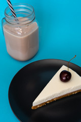 A glass mug of coffee with the tube and a piece of vanilla cheesecake with a cherry on a plate on a blue background. Close-up. Vertical. Top-side view