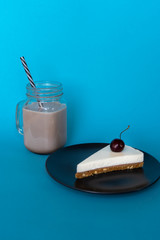 A glass mug of coffee with the tube and a piece of vanilla cheesecake with a cherry on a plate on a blue background. Vertical. 