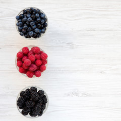 Bowls containing berries (blueberries, blackberries, raspberries) on a white wooden background, top view. Healthy eating and dieting. From above, overhead. Copy space.