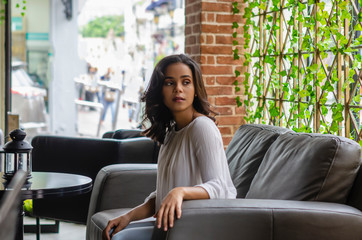 young girl from 19 to 25 years old sitting on black sofa in restaurant looking out the window waiting for her partner