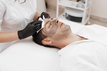 Obraz na płótnie Canvas Beautician applying cosmetic mask on man's face in spa salon.