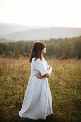 Fototapeta na wymiar Stylish boho girl walking in grass and wildflowers in sunny meadow at atmospheric sunset. Happy hipster woman in linen rustic dress enjoying traveling in evening mountains. Space for text