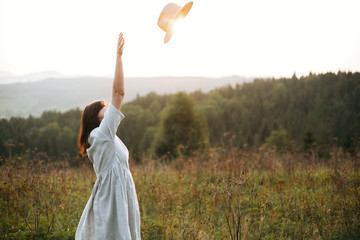 Stylish carefree boho girl throwing her hat in the sky in sunny light  at atmospheric sunset. Happy...