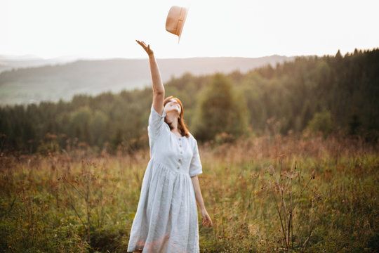 Stylish carefree boho girl throwing her hat in the sky in sunny light  at atmospheric sunset. Happy hipster woman in linen rustic dress enjoying traveling in evening  meadow