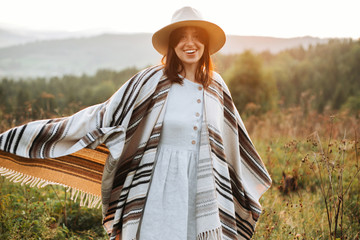 Stylish boho girl smiling  in sunny light at atmospheric sunset in meadow. Happy hipster woman in...