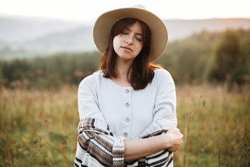 Stylish boho girl portrait in sunny light at atmospheric sunset in meadow. Calm hipster woman in poncho and hat enjoying traveling in evening mountains. Space for text.