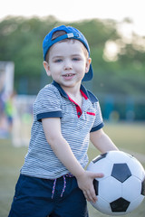 Portrait of little toddler child standing in green football field holding soccer ball. Smiling little football player at stadium. Future footbal star and active childhood concept